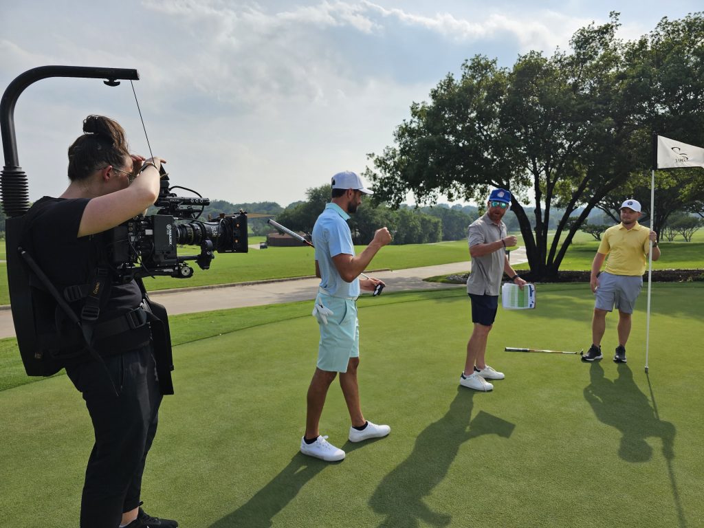 Woman with camera filming two golfers and a director on a golf course